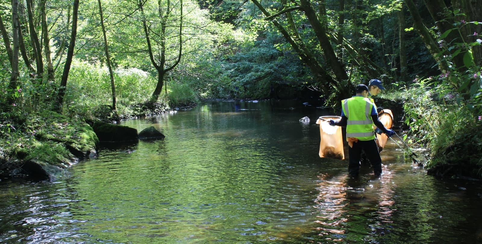 Two people collect trash in a river. They wear vests, carry a trash bag, and hold a trash scooper.