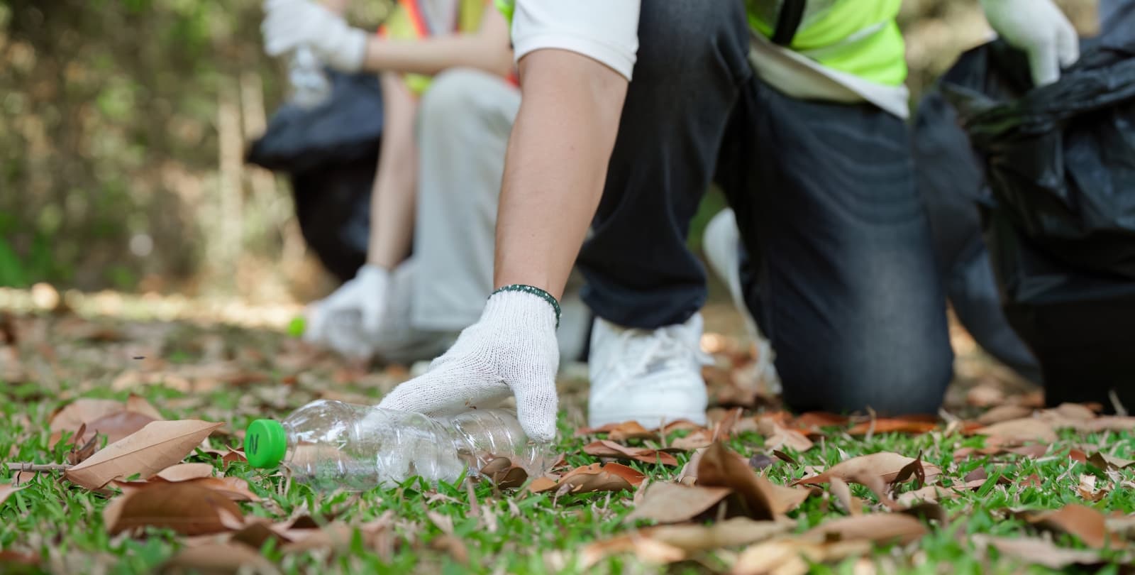 A person rests on one knee to pick up a plastic water bottle off the grass.