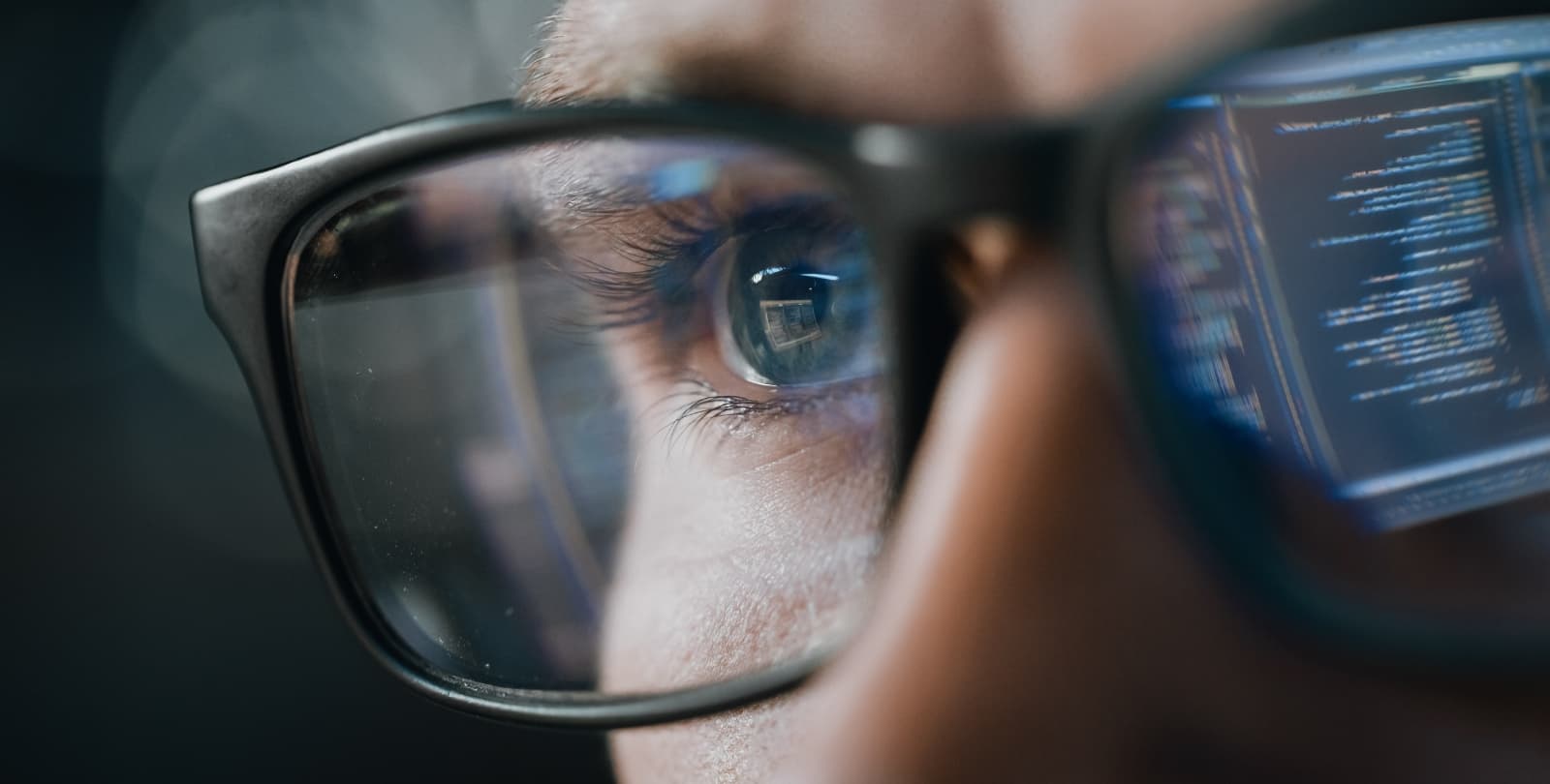 Close-up photo of a man looking at a computer screen. He is wearing glasses, and in the screen's reflection is in his left lens.