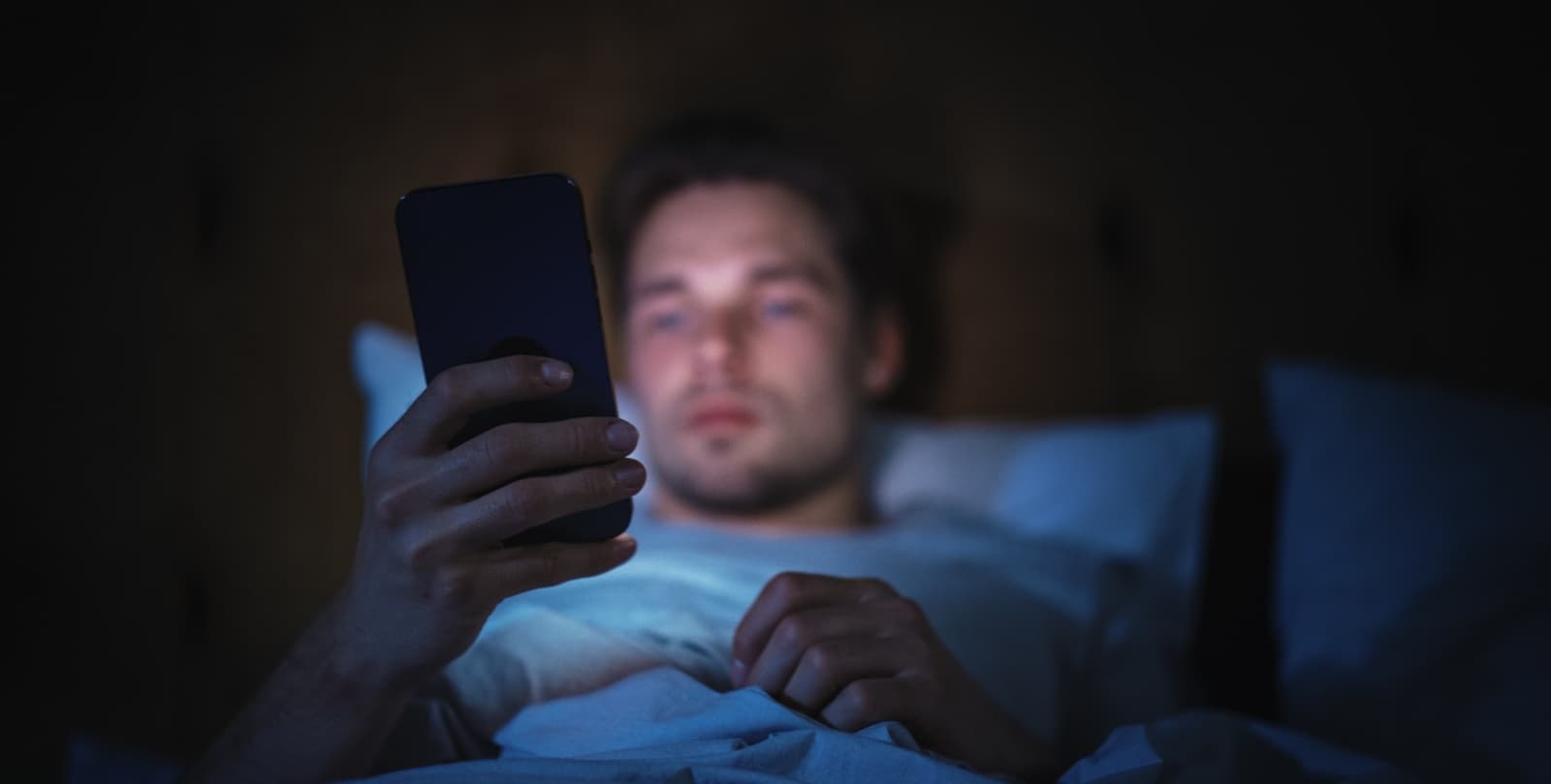 A young man uses his phone while laying in bed in a dark room. His face is illuminated by the screen.