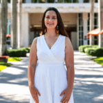 A photo of Rachel Mutti. Rachel has long dark hair, is wearing a white dress, and is standing outside on a walkway on a college campus.