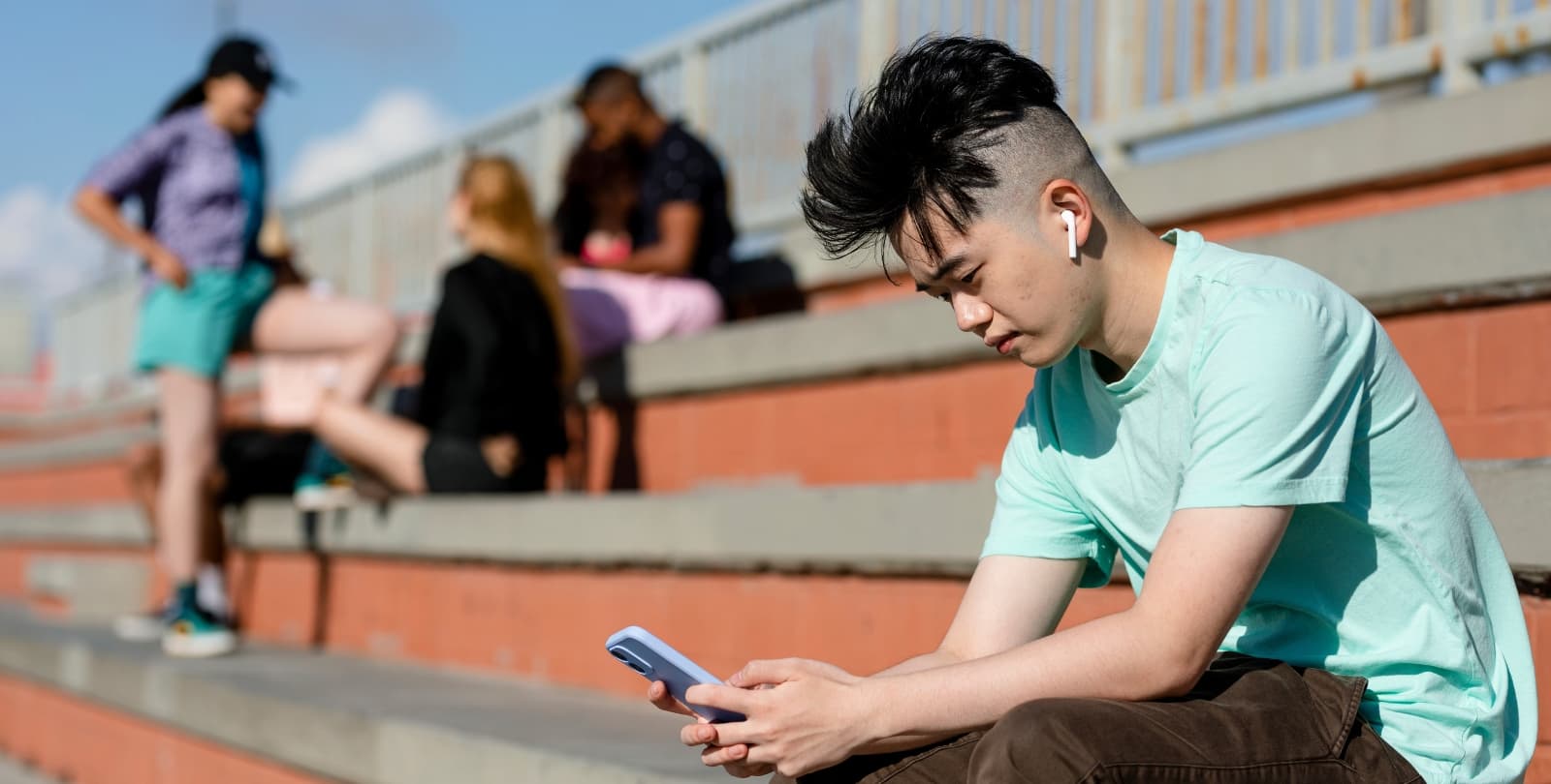 A teen sits alone on the bleachers, phone in hand and AirPods in his ears. A group of teens talks on the other side of the bleachers.