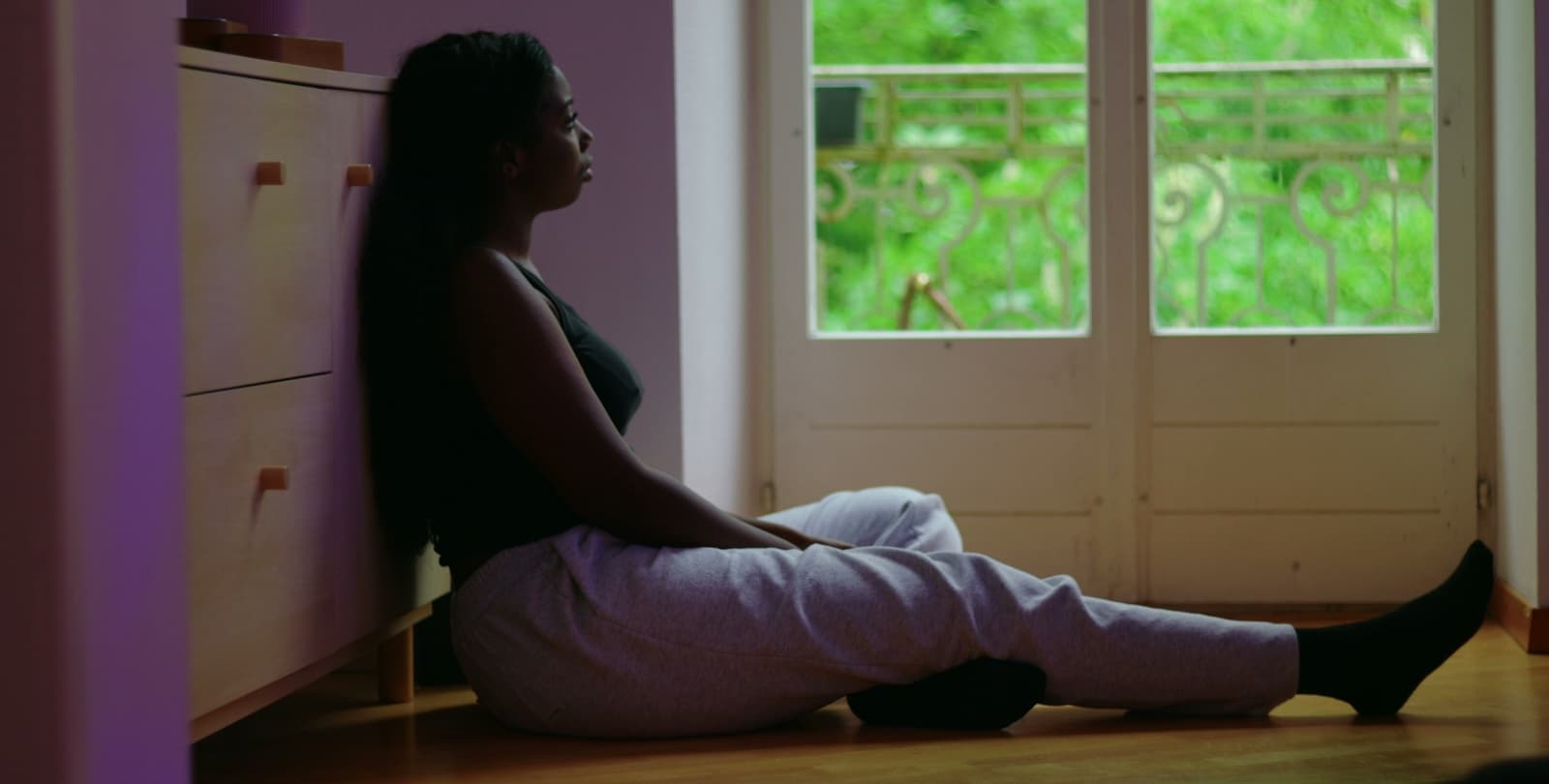 A woman sits on the ground, her back leaned against a dresser. She looks sadly out the window.