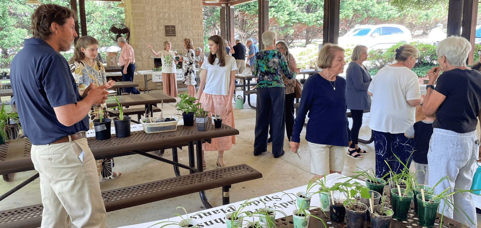A group of people stand and talk around tables in a park shelter. Various small plants sit on the tables.