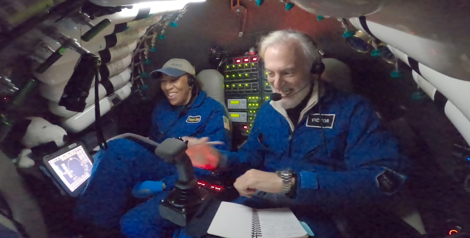 Dr. Dawn Wright and CDR Victor Vescovo sit in the submersible DSV Limiting Factor during a dive. Both wear blue flight suits; there is a thumbstick in front of Victor, and a panel of lighted buttons behind them.