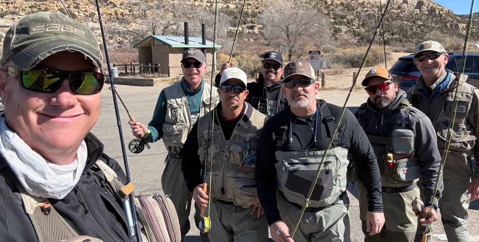 A group of seven men wear waders and hold fishing poles on a sunny day.