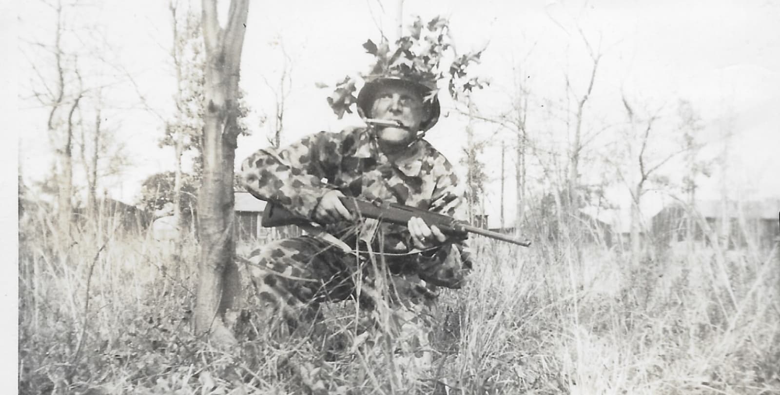 Black and white photo of Staff Sergeant William Black. Black is wearing camo, holding a rifle, and crouching in tall grass.