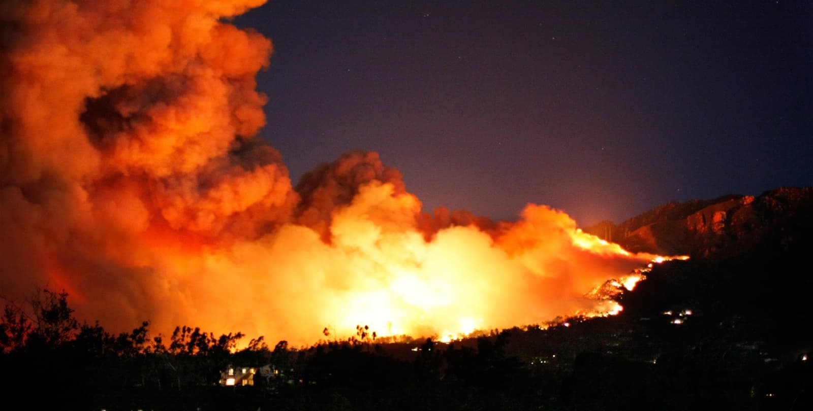 Photo of the 2008 Tea Fire. A massive, orange cloud of smoke illuminates the night sky as flames race toward a series of houses.