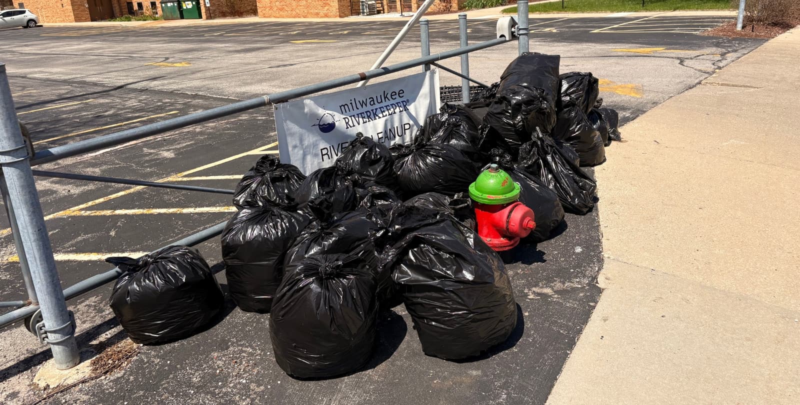 A pile of twenty full garbage bags sit in front of a sign that reads "Milwaukee Riverkeeper River Cleanup"