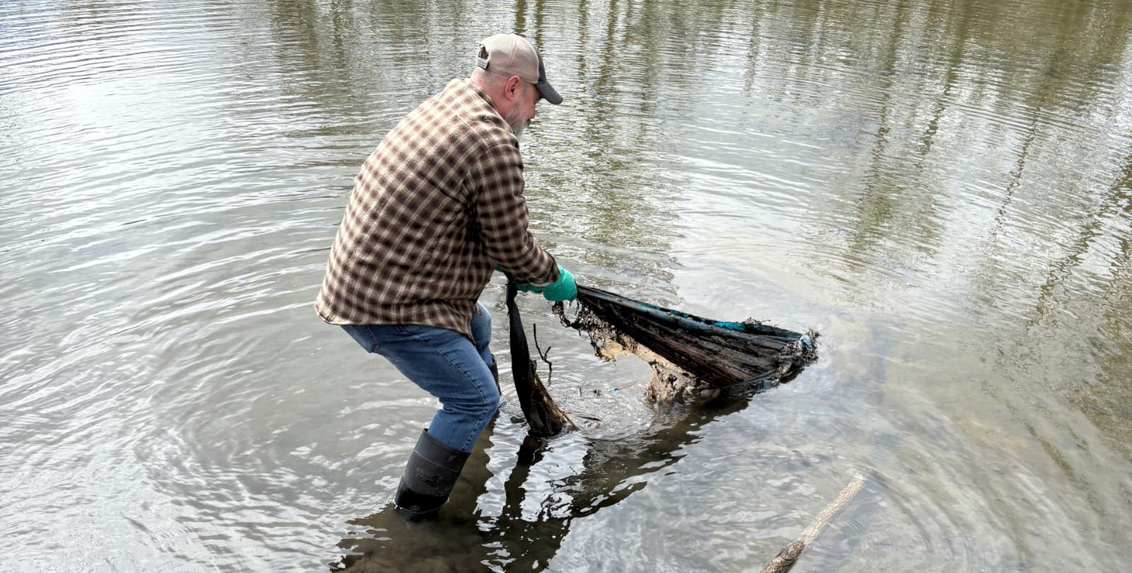 A man wearing a baseball hat and flannel shirt pulls a large piece of trash, which appears to be mesh and metal, out of Lincoln Creek.