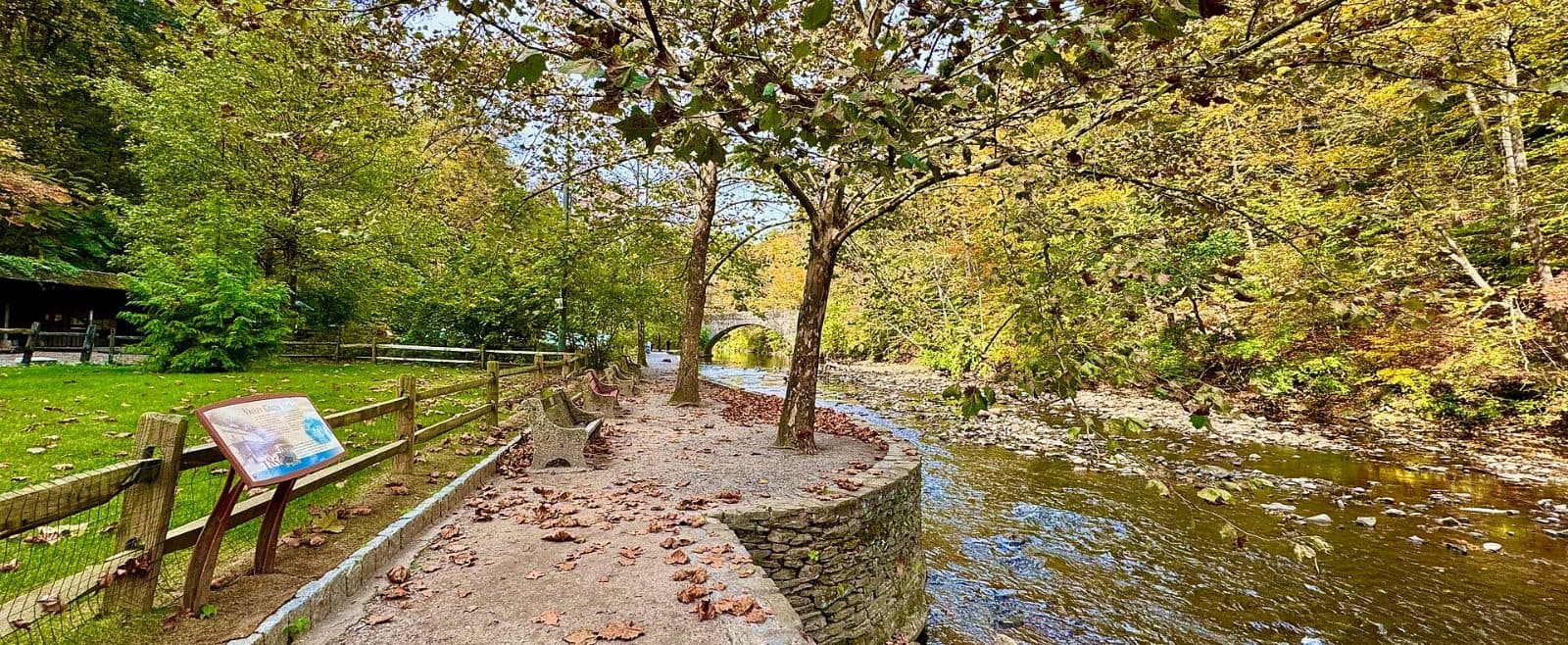 A winding stone path directly next to a creek. A fence runs along the left side, and benches face the creek.